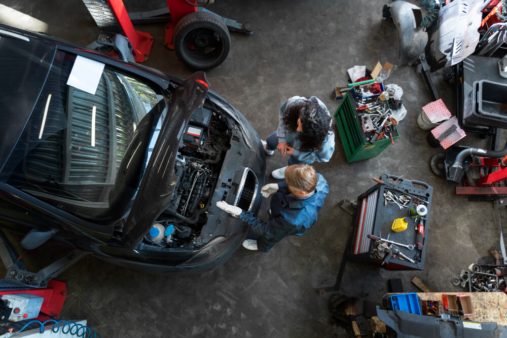 Zwei Personen prüfen den Motor eines Autos in einer Werkstatt, während Werkzeuge und Ausrüstung um sie herum verstreut sind