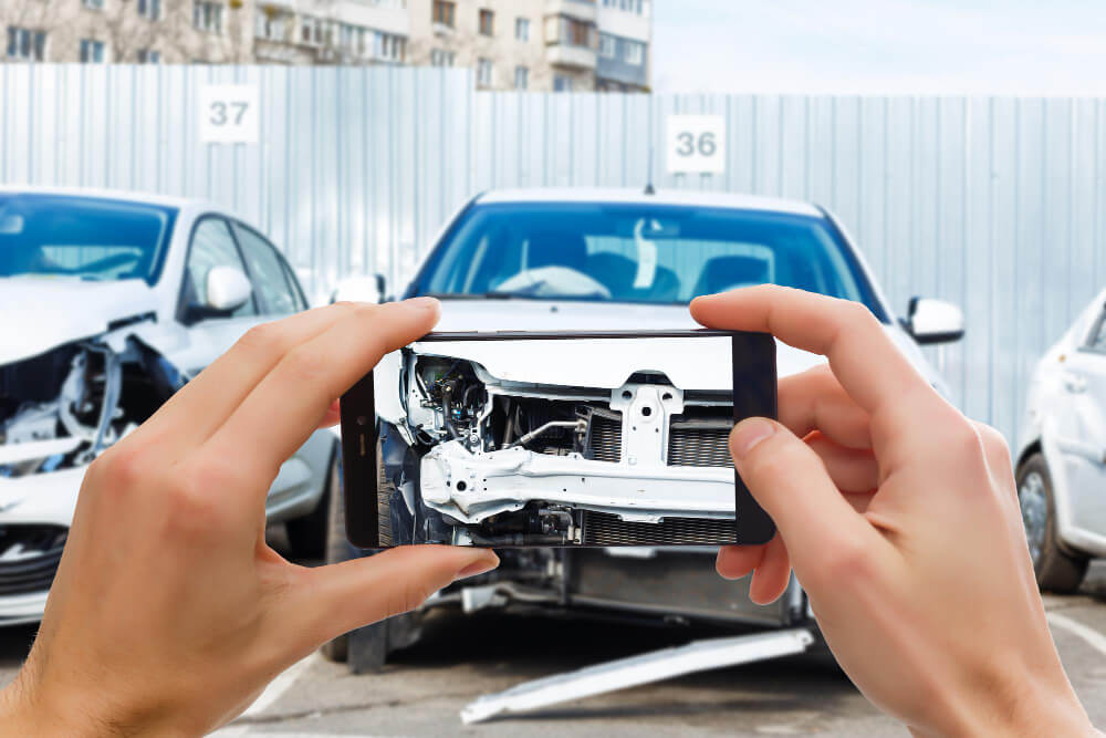 Eine Person fotografiert mit dem Smartphone ein beschädigtes Auto, im Hintergrund stehen zwei Unfallfahrzeuge auf einem Parkplatz
