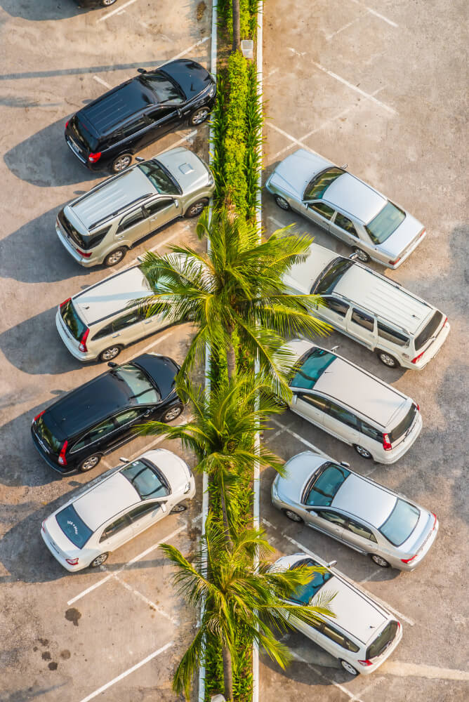 Aus der Vogelperspektive sind Autos schräg zu beiden Seiten einer Palmenreihe auf einem Parkplatz geparkt