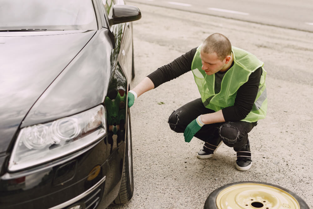 Ein Mann mit grüner Warnweste und Handschuhen hockt am Auto und prüft einen Reifen, daneben liegt ein Ersatzrad auf dem Boden