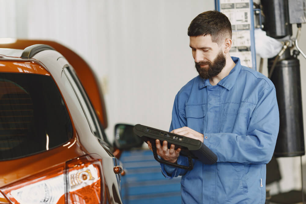 Ein Mechaniker in blauer Arbeitskleidung bedient ein Diagnosegerät neben einem orangefarbenen Auto in einer Werkstatt