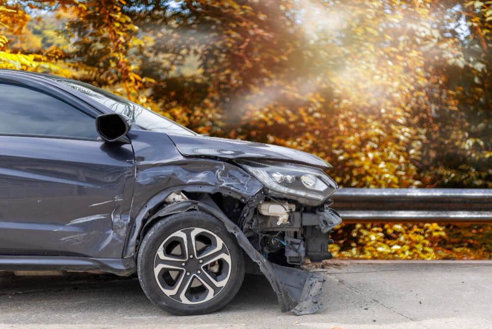 Ein graues Auto mit beschädigter Front und fehlender Stoßstange steht an einer Leitplanke, im Hintergrund sind herbstliche Bäume zu sehen